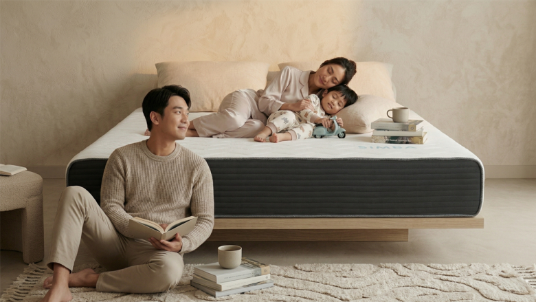 奶凍捲床墊居家生活場景：一對年輕夫妻與小孩在日系簡約風格的臥室中。女性與小孩舒適地躺在具有深灰色護邊與白色波浪面層的床墊上，男性則坐在床邊地毯閱讀。畫面傳遞出護脊撐腰、穩定且安心的睡眠環境氛圍。