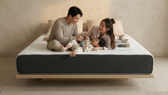 這張圖片的主題是「奶凍捲床墊」,展示了一個亞洲家庭在床上溫馨互動的畫面。 一對父母和他們的年幼兒子坐在一張淺色木質懸浮床架上的床墊上。床墊表面為白色,側邊則是深灰色的布料,呈現出如奶凍捲般整齊分層的視覺效果。 父親身穿灰色毛衣,母親穿著粉色睡衣,兒子則穿著印有動物圖案的連身衣。父親笑著看著兒子,兒子則躺在母親懷裡,手裡拿著一台玩具飛機。母親開心地笑著,轉頭看向兒子。 床墊後方擺放著幾顆米色枕頭。右側床頭櫃上擺放著一疊書和一個白色馬克杯。背景牆面呈現米白色,帶有自然的紋理。 整個畫面呈現溫暖、舒適和清新的氛圍。